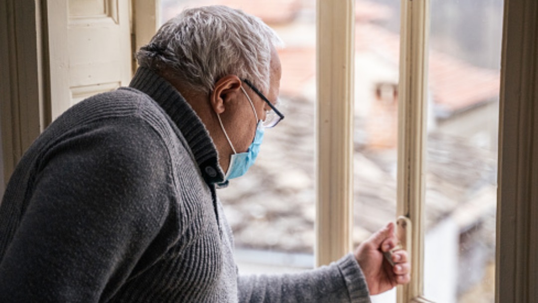 Un abuelito usa cubrebocas mientras ve por la ventana