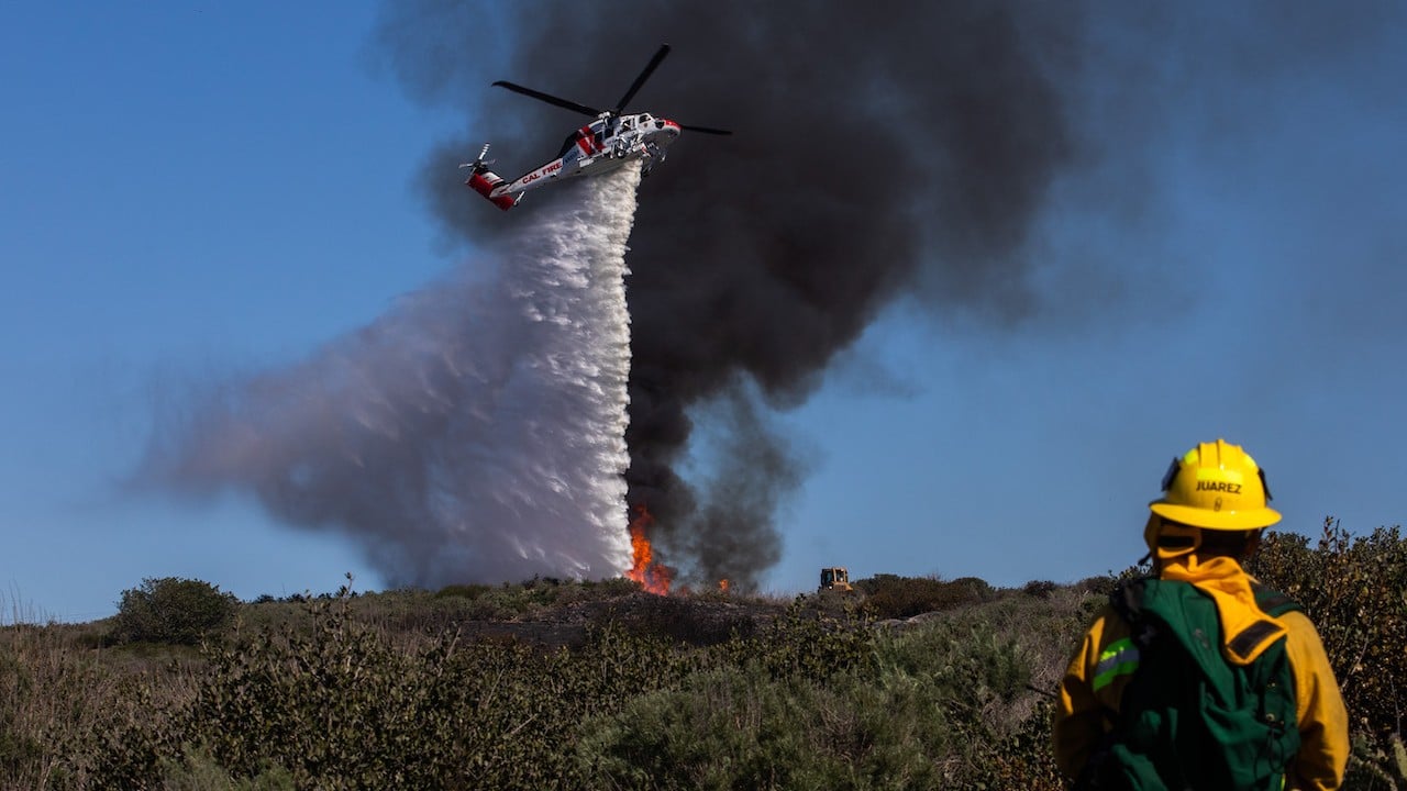 Un bombero observa cómo un helicóptero arroja agua sobre una ladera humeante en Laguna Beach, California, 10 de febrero de 2022 (Getty Images)